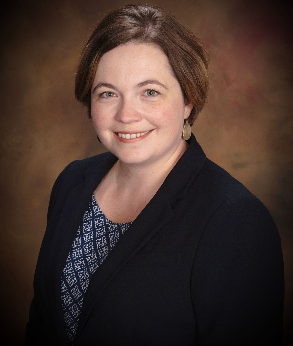 Woman in a dark blazer smiling, with a patterned blouse, against a blurred brown background.