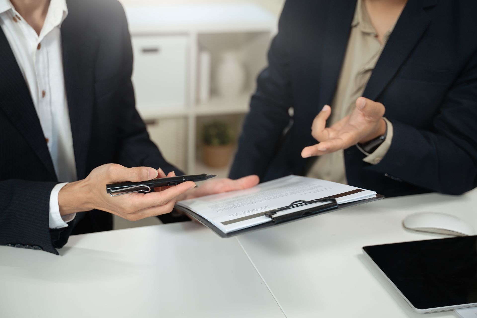 Two people in suits at a desk reviewing documents; one holds a pen, gesturing.