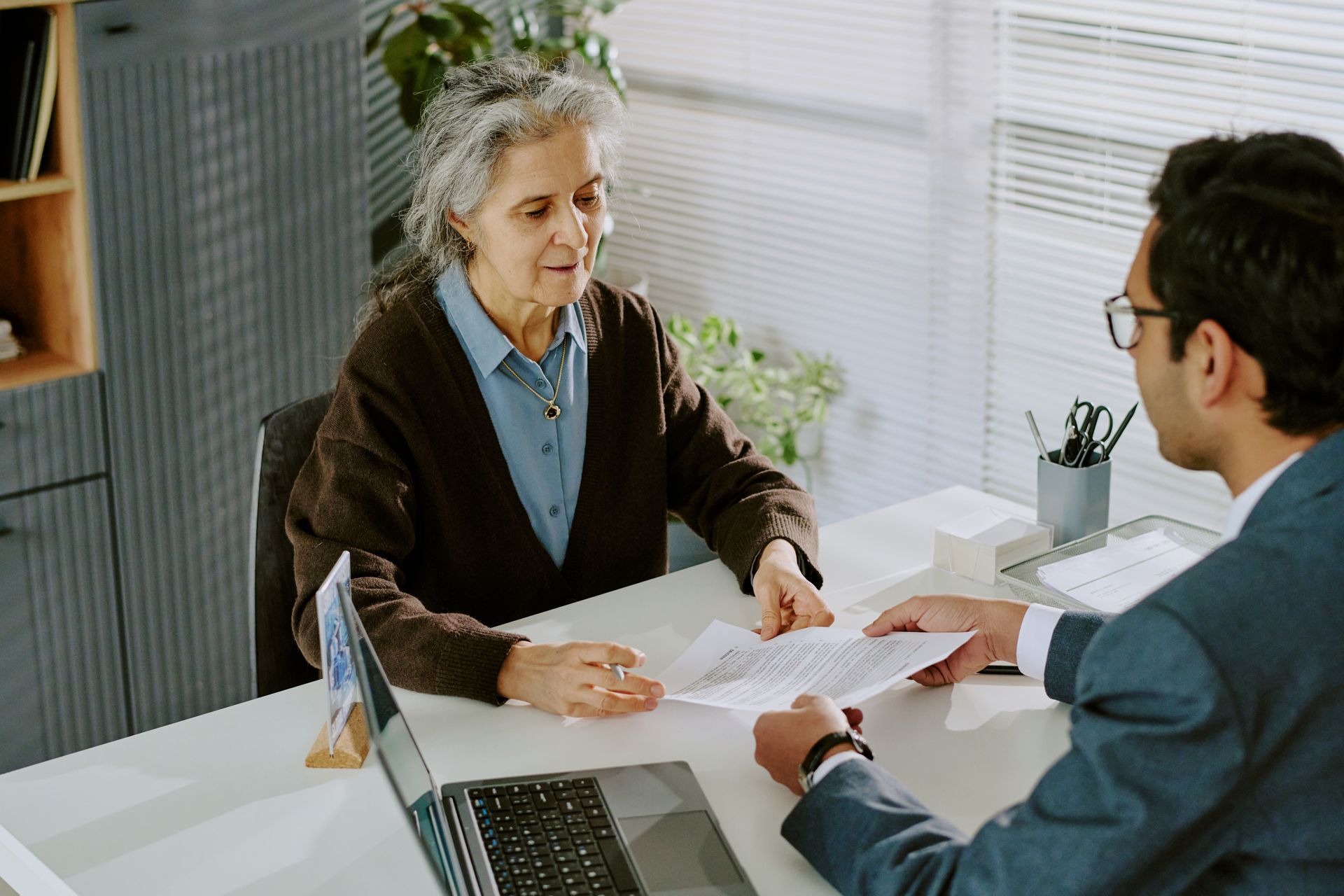 An older person reviews documents with a person in a suit at a desk in an office.