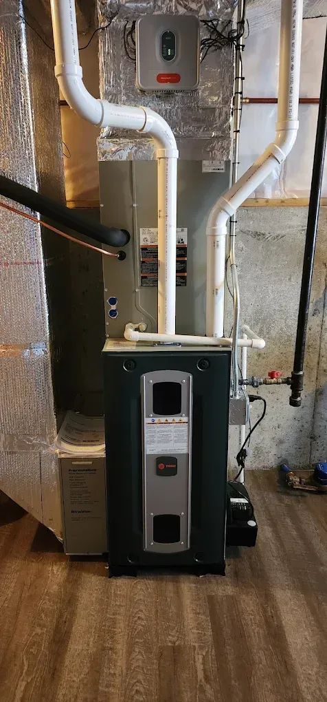 A furnace and water heater are in a basement. White PVC pipes are connected to the furnace. The area has wood-look flooring.