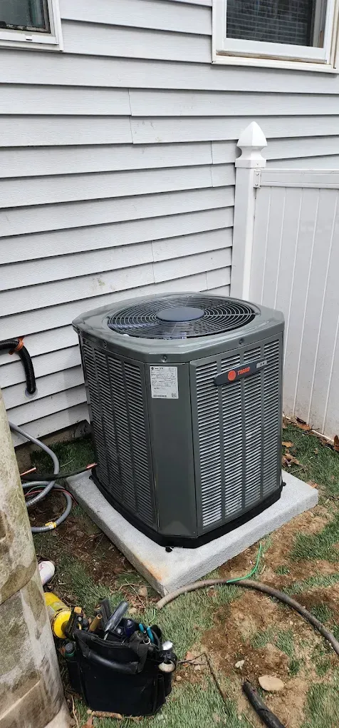 An air conditioning unit on a concrete pad next to a house with white siding. Tools on the ground.