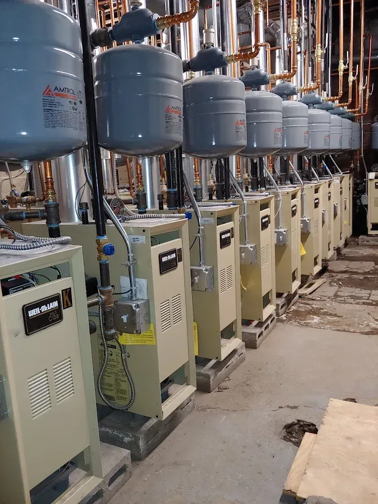 Row of industrial water heaters with attached tanks and piping, indoors.
