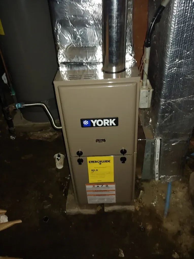 York furnace in a basement. Beige front with blue logo, silver flue, surrounded by ductwork and plumbing.