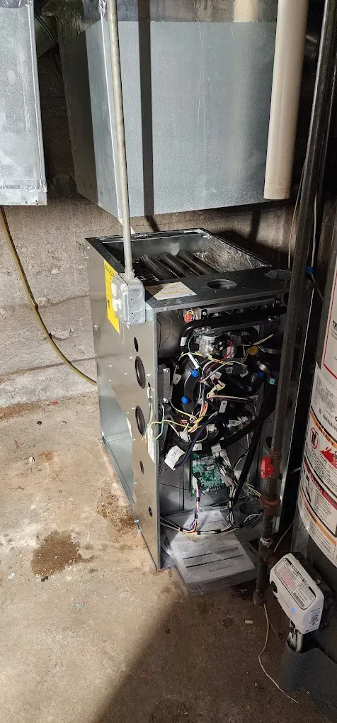 Furnace with open panels, showing internal components. The unit sits on a concrete floor near a water heater.