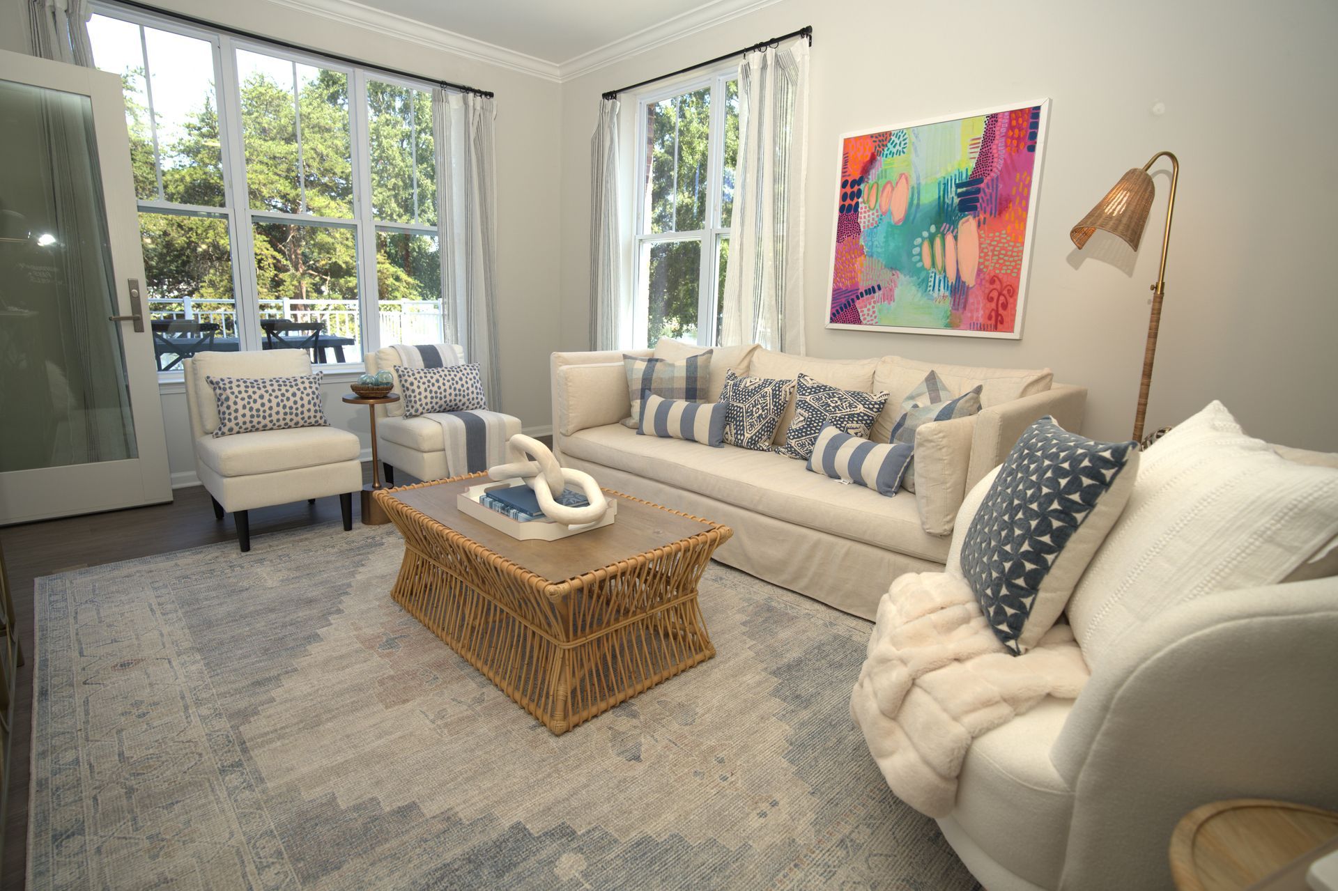 Living room with white furniture, blue accents, rattan coffee table, and colorful abstract art.
