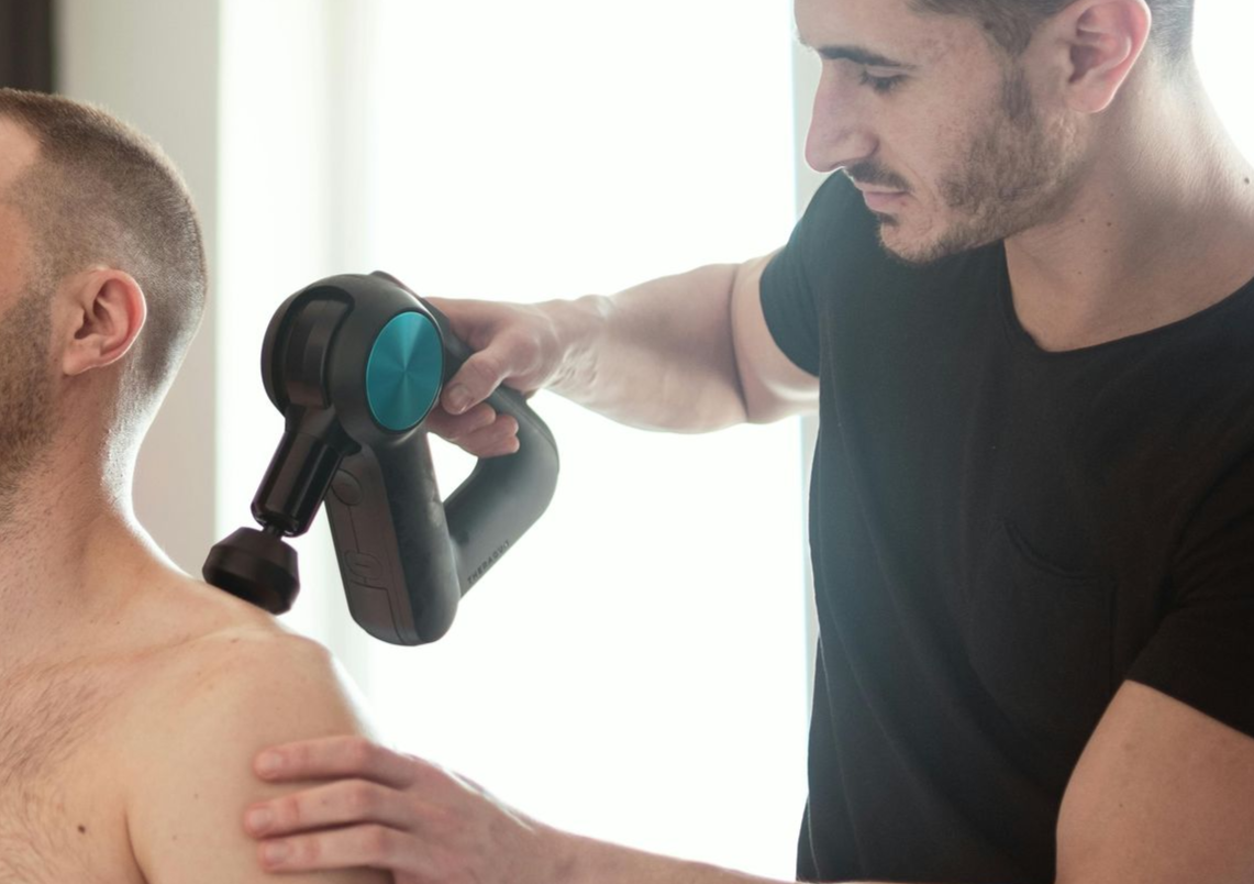 Man using a massage gun on another person’s shoulder in a bright room