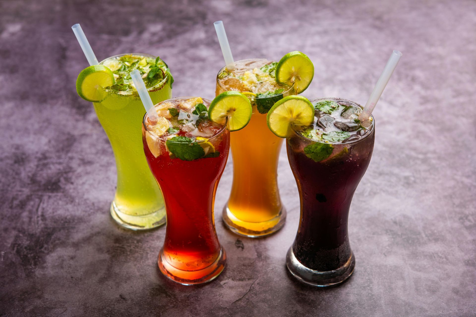 Four colorful tropical cocktails with lime garnishes and straws on a gray surface