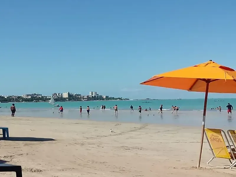 Cena de praia com guarda-sol laranja, pessoas na água e prédios ao fundo sob um céu azul.