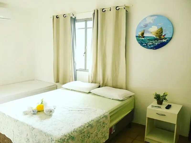 Quarto com uma cama de casal, uma cama de solteiro e uma pequena mesa de cabeceira. Paredes e cortinas em tons claros com uma pintura do oceano.