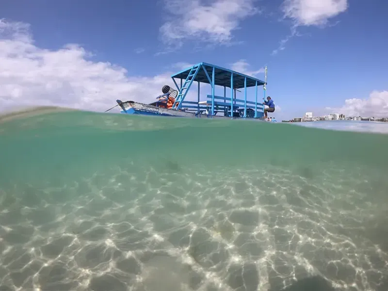 Vista dividida de uma jangada azul em águas cristalinas. Pessoas sentadas na jangada sob um dossel azul, com a cidade visível no horizonte.