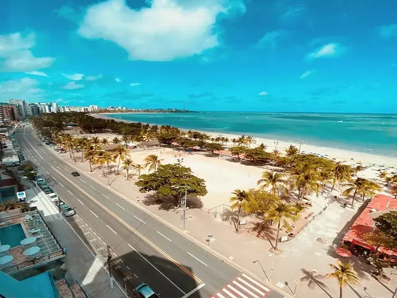 Paisagem costeira com estrada, praia, palmeiras, edifícios e céu azul.