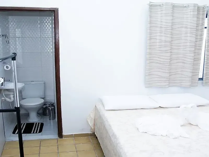 Quarto com cama, paredes brancas e banheiro visível através de uma porta.
