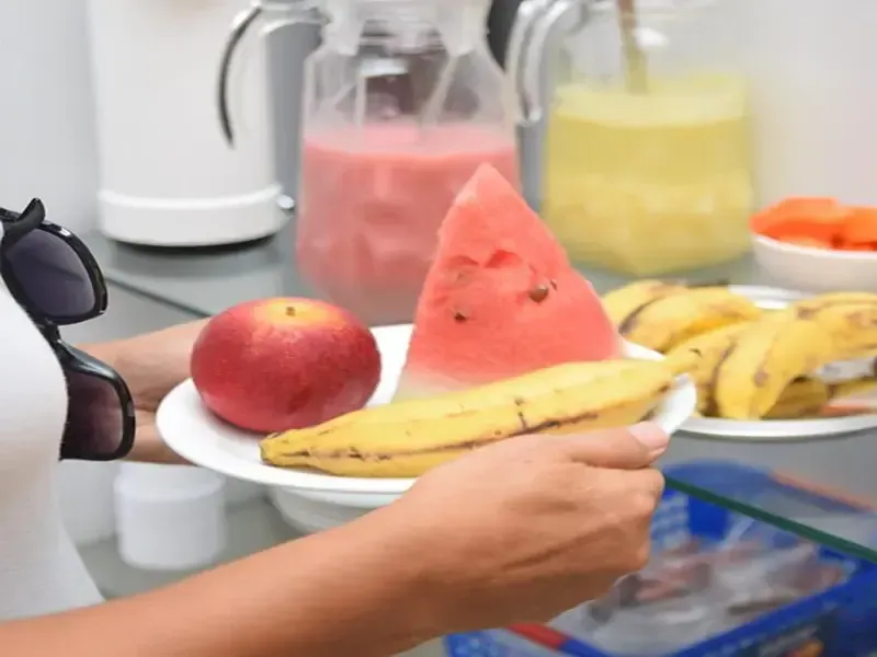 Mulher segurando um prato de frutas: maçã, fatia de melancia e banana. Sucos ao fundo.