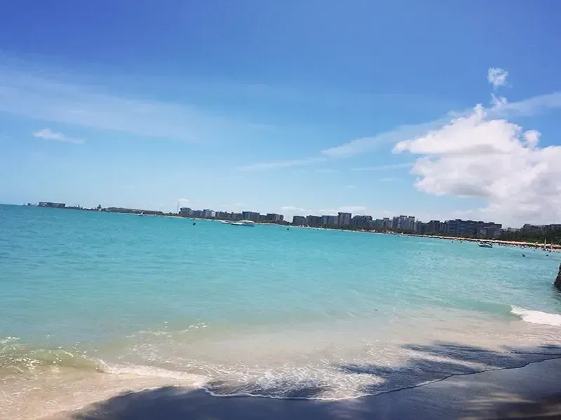 Águas turquesas encontram a praia de areia sob um céu azul, com edifícios ao longe.