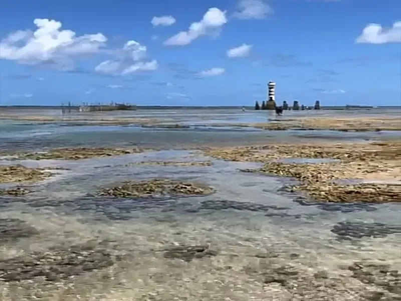 Paisagem costeira com águas rasas e rochosas e um farol sob um céu azul com nuvens.