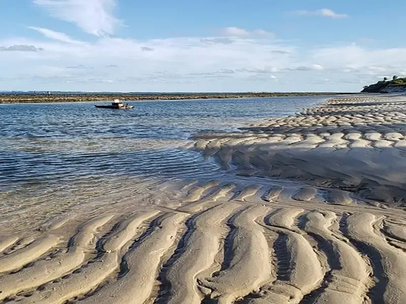Praia de areia com sulcos cheios de água, que levam a um mar calmo e a um barco ao longe sob um céu azul.