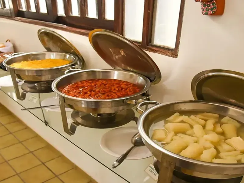 Buffet com rechauds contendo alimentos amarelos, vermelhos e brancos, perto de uma janela.