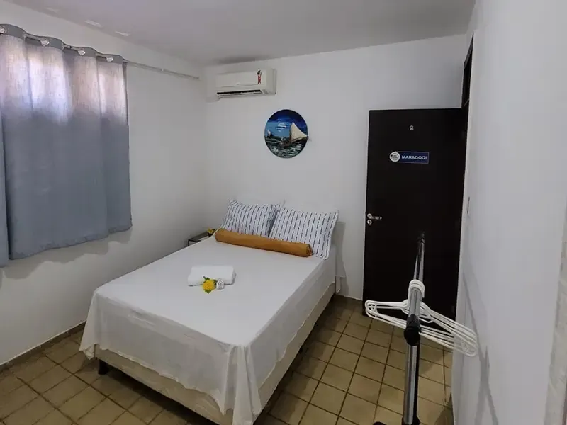 Quarto pequeno com cama, janela com cortinas e porta escura; inclui ar condicionado.