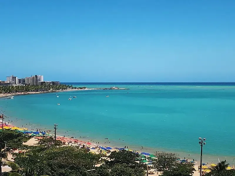 Cena de praia: água turquesa, céu azul, pessoas na areia, guarda-sóis, prédios ao longe.