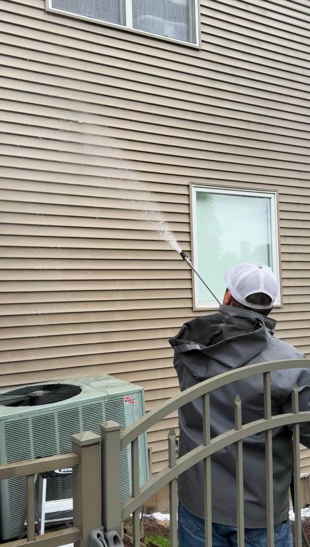 A person in a blue shirt and cap uses a hose to spray cleaning foam on the dirty exterior wall of a white house.