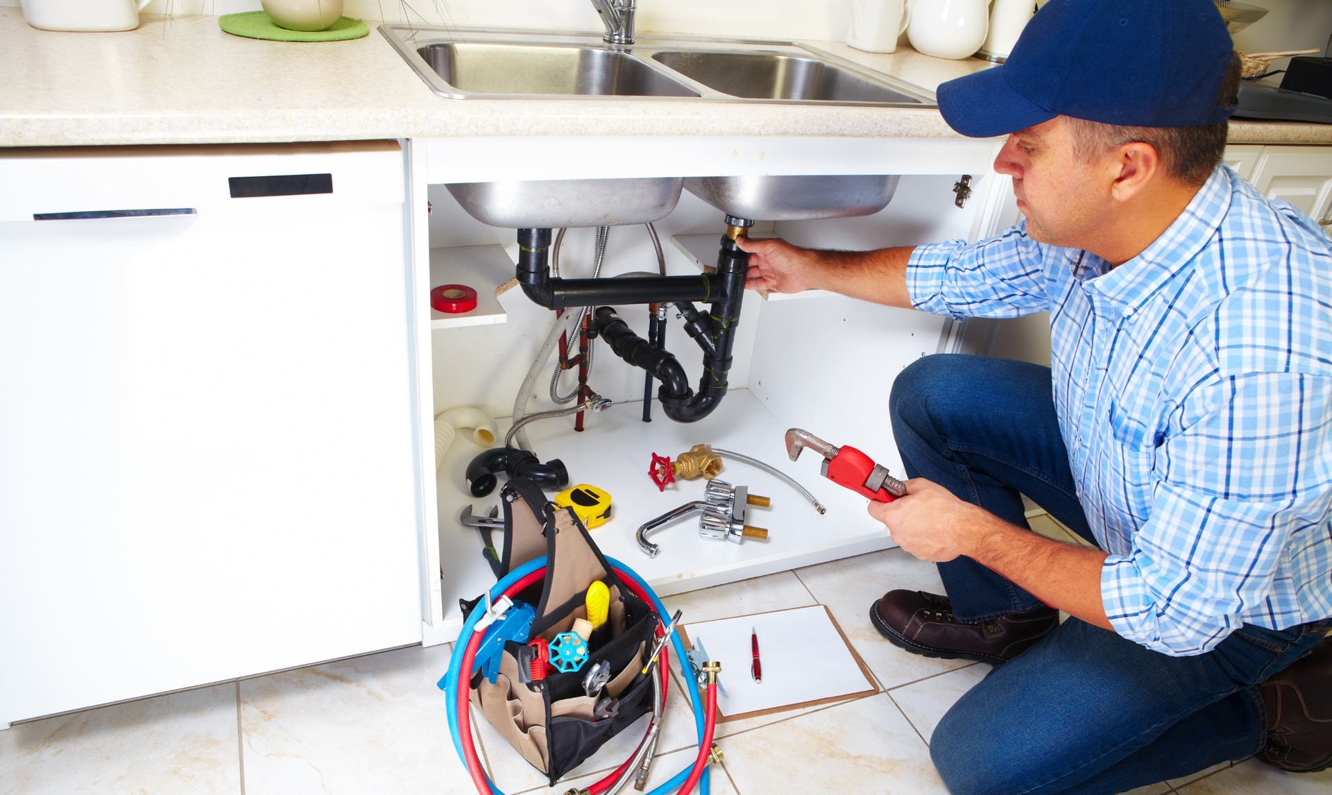 Plumber in a blue cap and plaid shirt repairs sink pipes, kneeling with tools and a toolbox under the sink.