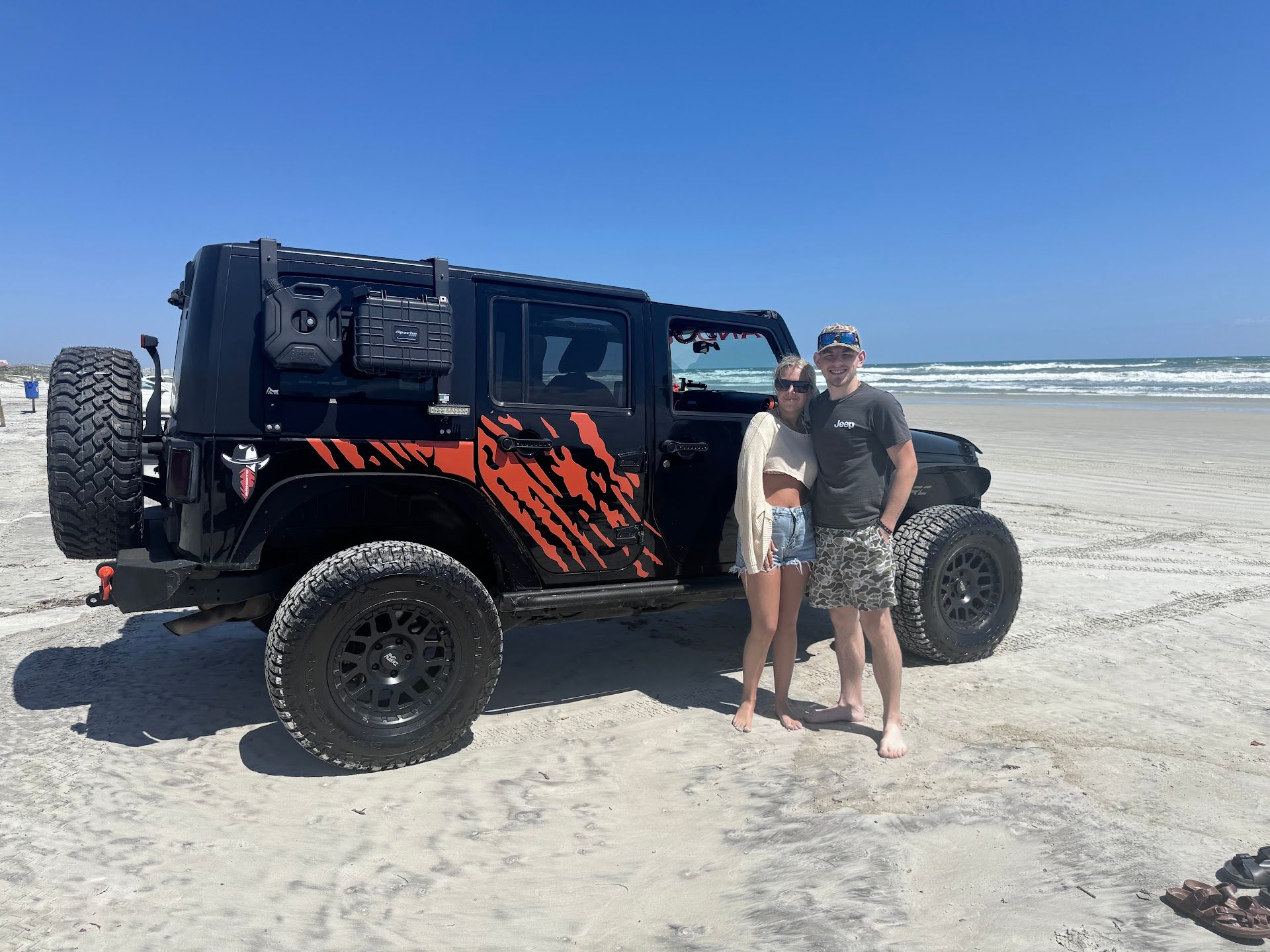 A couple stands next to a black Jeep on a beach with the ocean in the background | Daytona Automotive