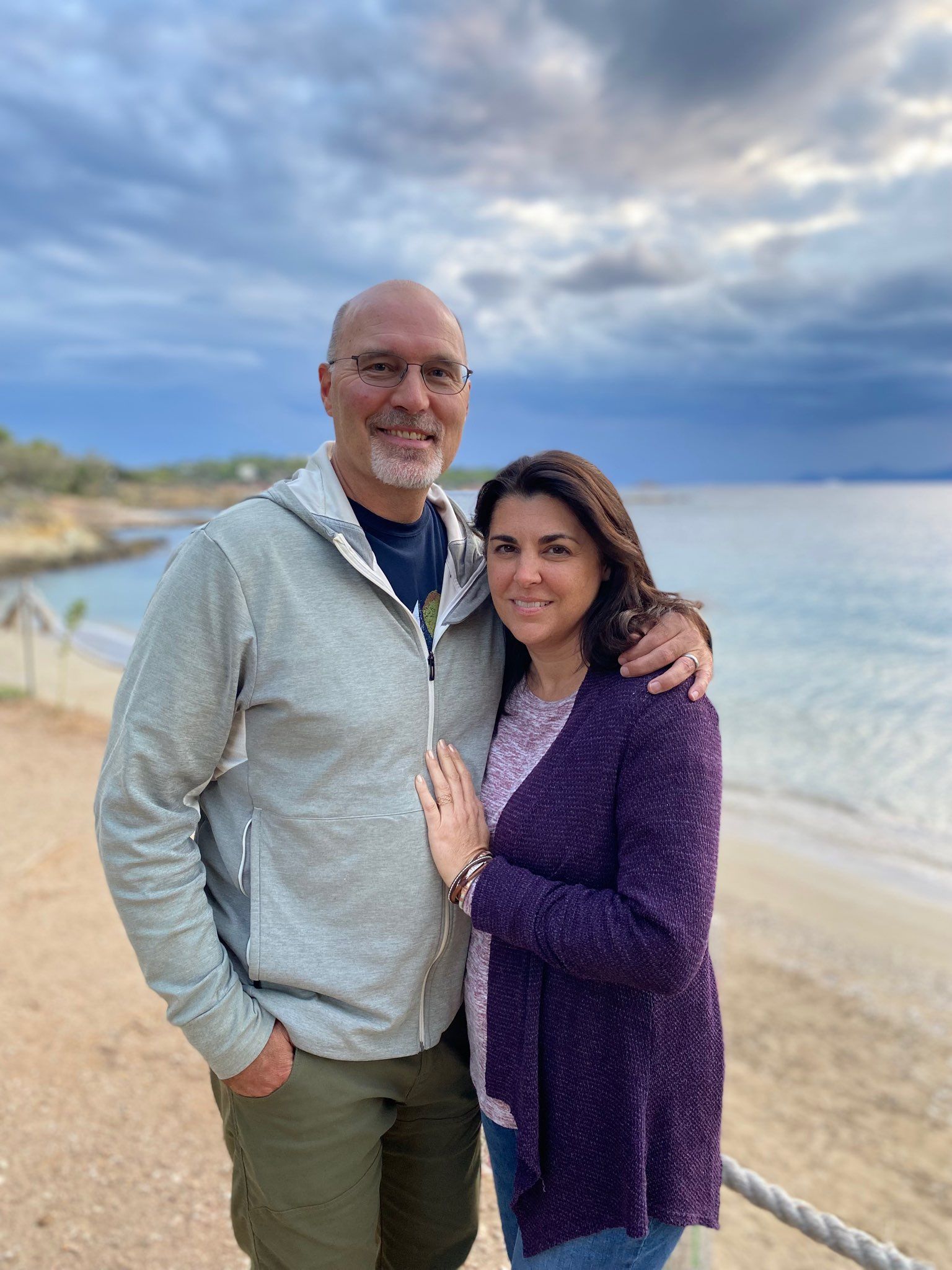 Couple embraces on a sandy beach. Man in gray jacket, woman in purple cardigan. Ocean and sky in background.