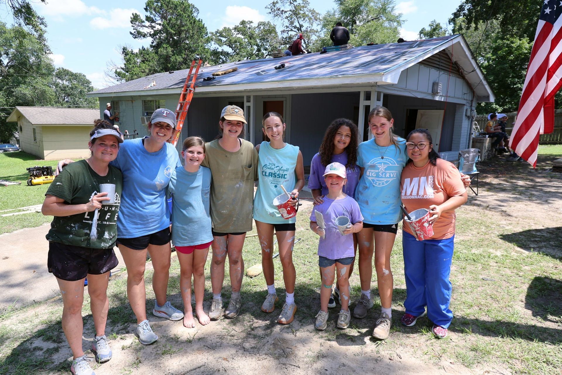 Group of people smiling in front of a house, some with snacks. People are working on the roof. American flag present.