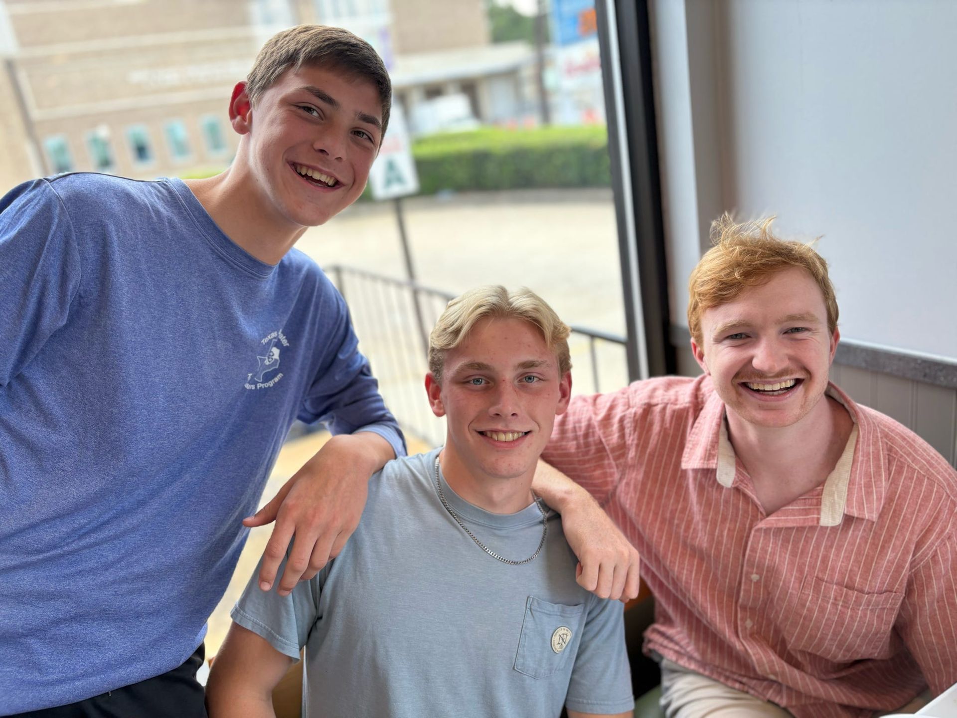 Three young people smiling for the camera; one with arm around another. Outdoors seen through window.