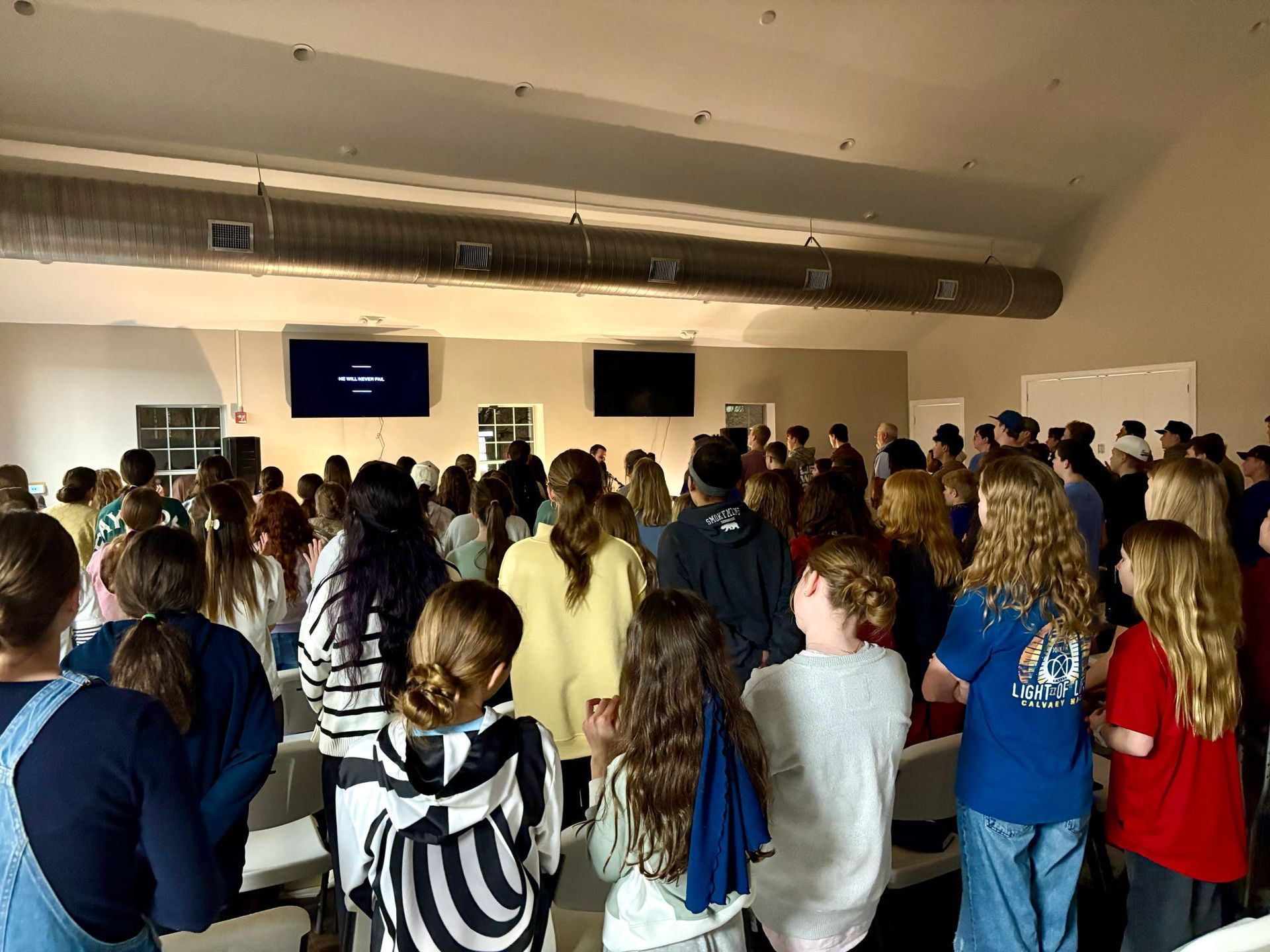 Large group of people gathered in a large room, looking towards two screens.