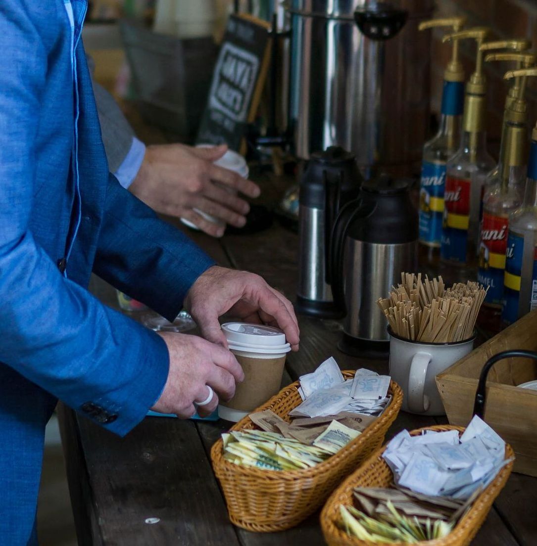 Person in blue suit holding a coffee cup at a coffee station with sugar packets and stirrers.