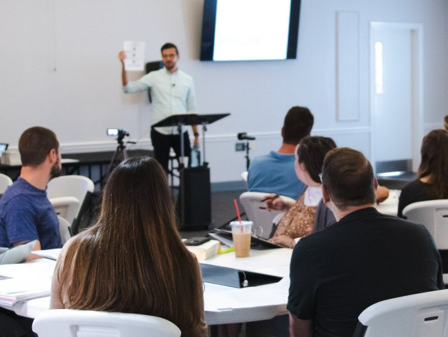 Man presenting to a group in a classroom setting, holding papers. Attendees at tables.