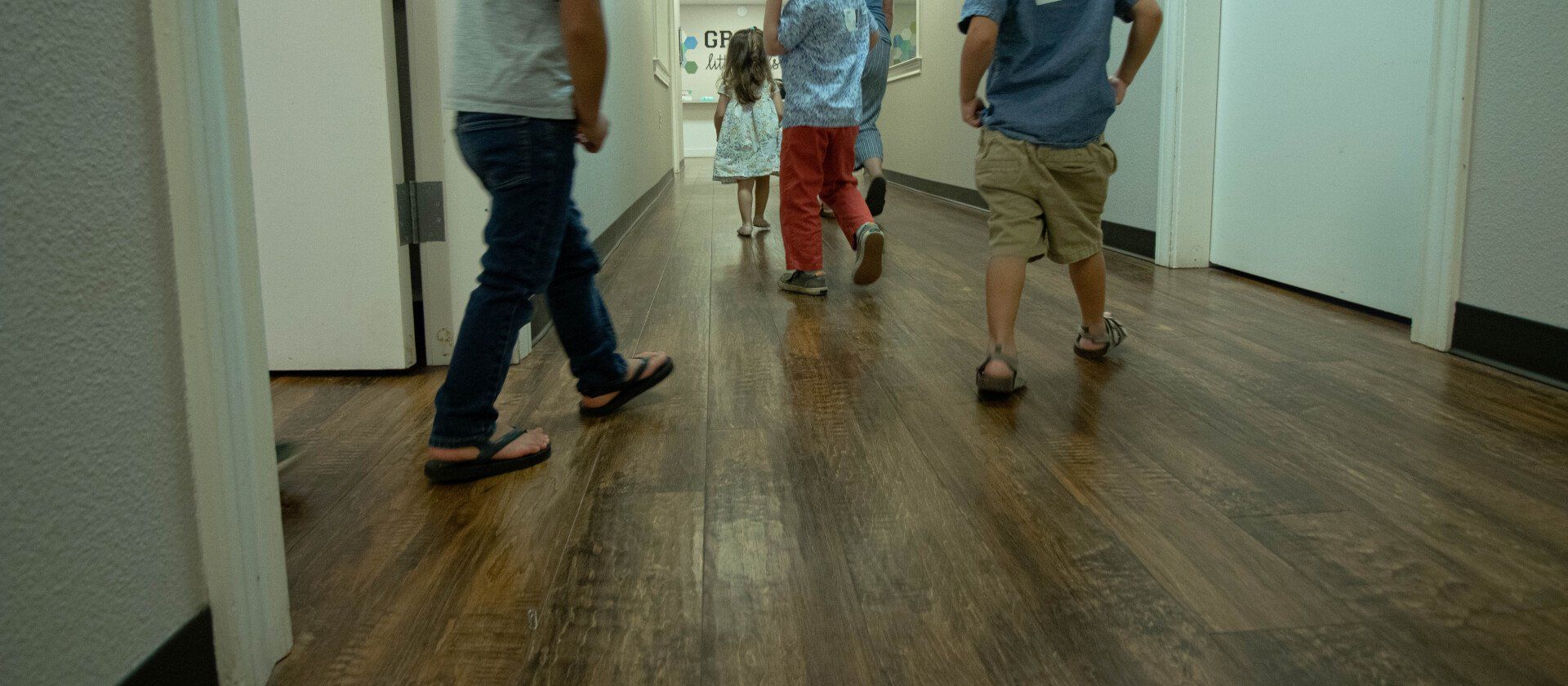 People walking down a wooden hallway; one person in jeans and sandals, others in shorts and shirts.