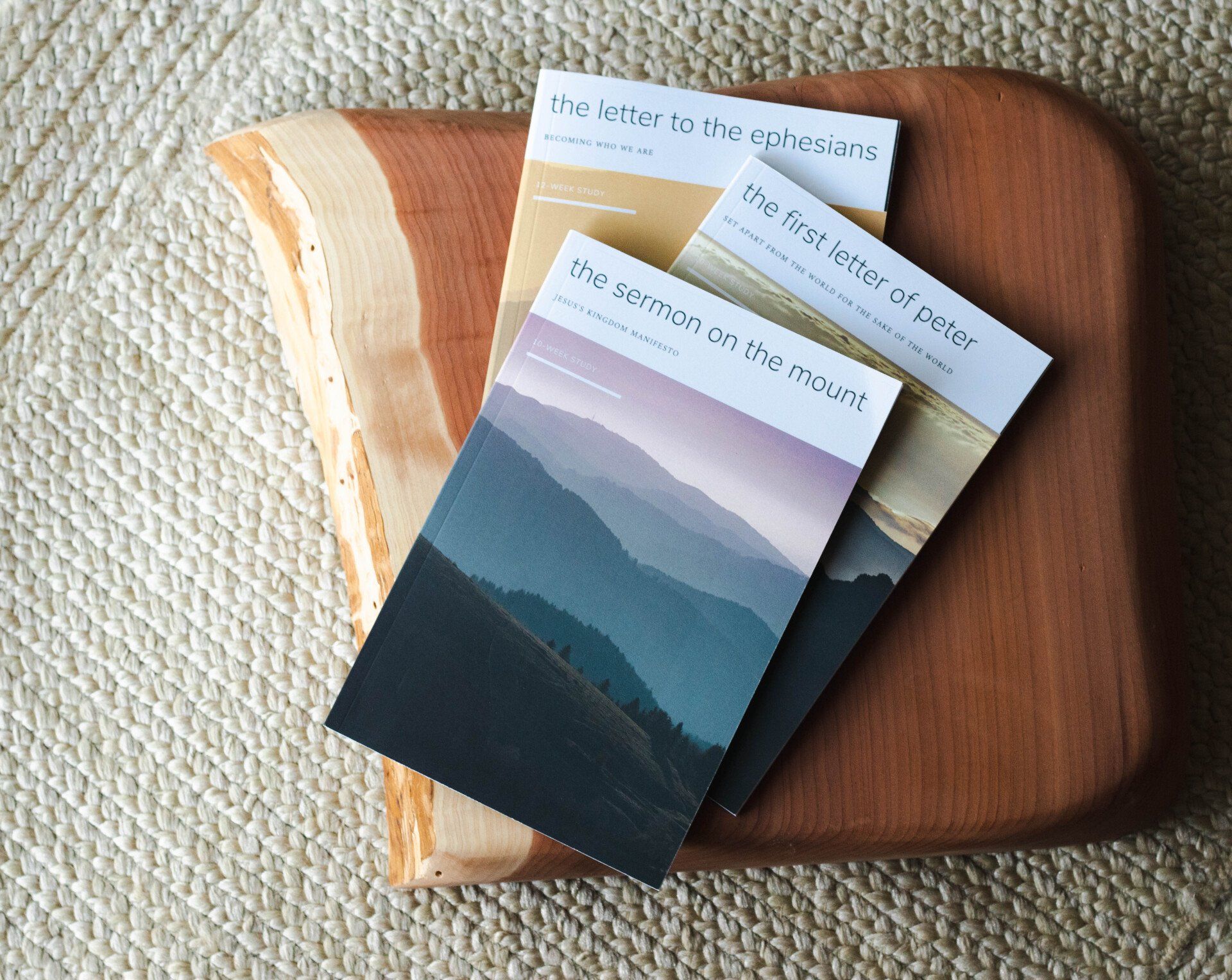 Three workbooks with mountain scenes stacked on a wooden cutting board against a tan woven surface.
