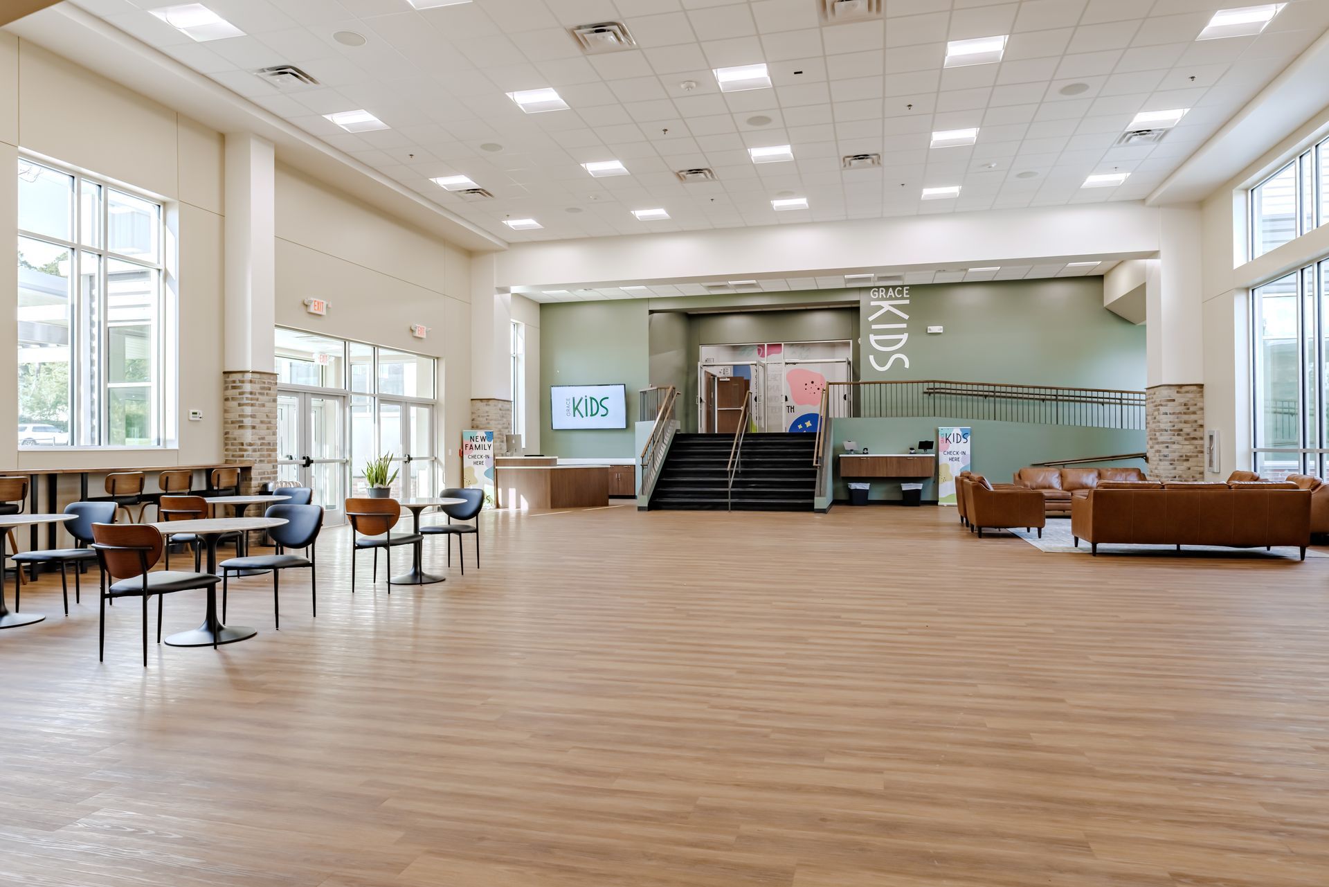 A spacious church lobby with light wood floors, tables, chairs, and couches leading to a set of stairs.