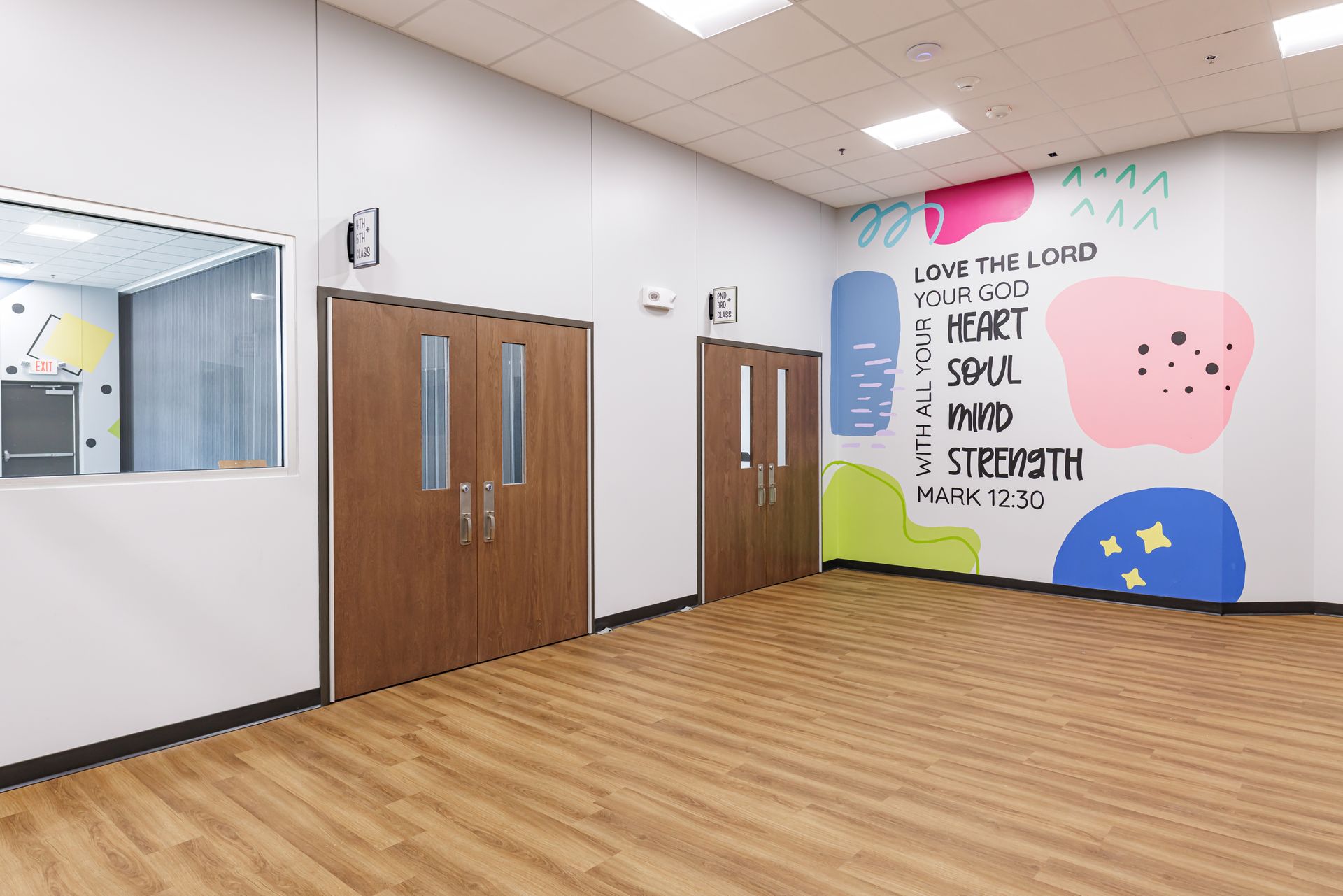 A bright hallway featuring two wooden doors and a wall mural with colorful shapes and text about loving the Lord.