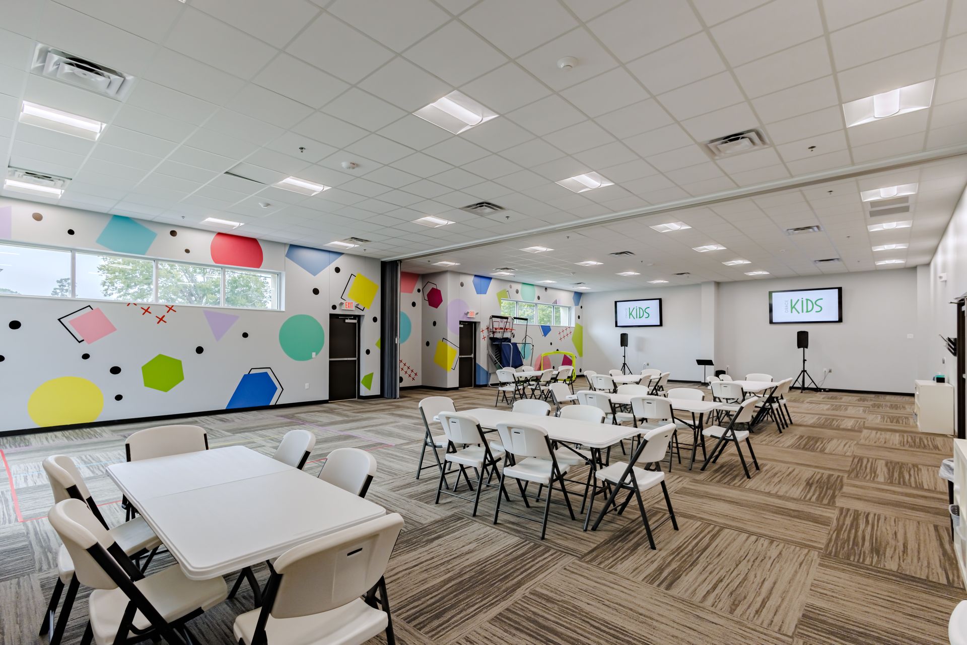 A large, brightly lit meeting room with tables, folding chairs, and colorful geometric wall art.