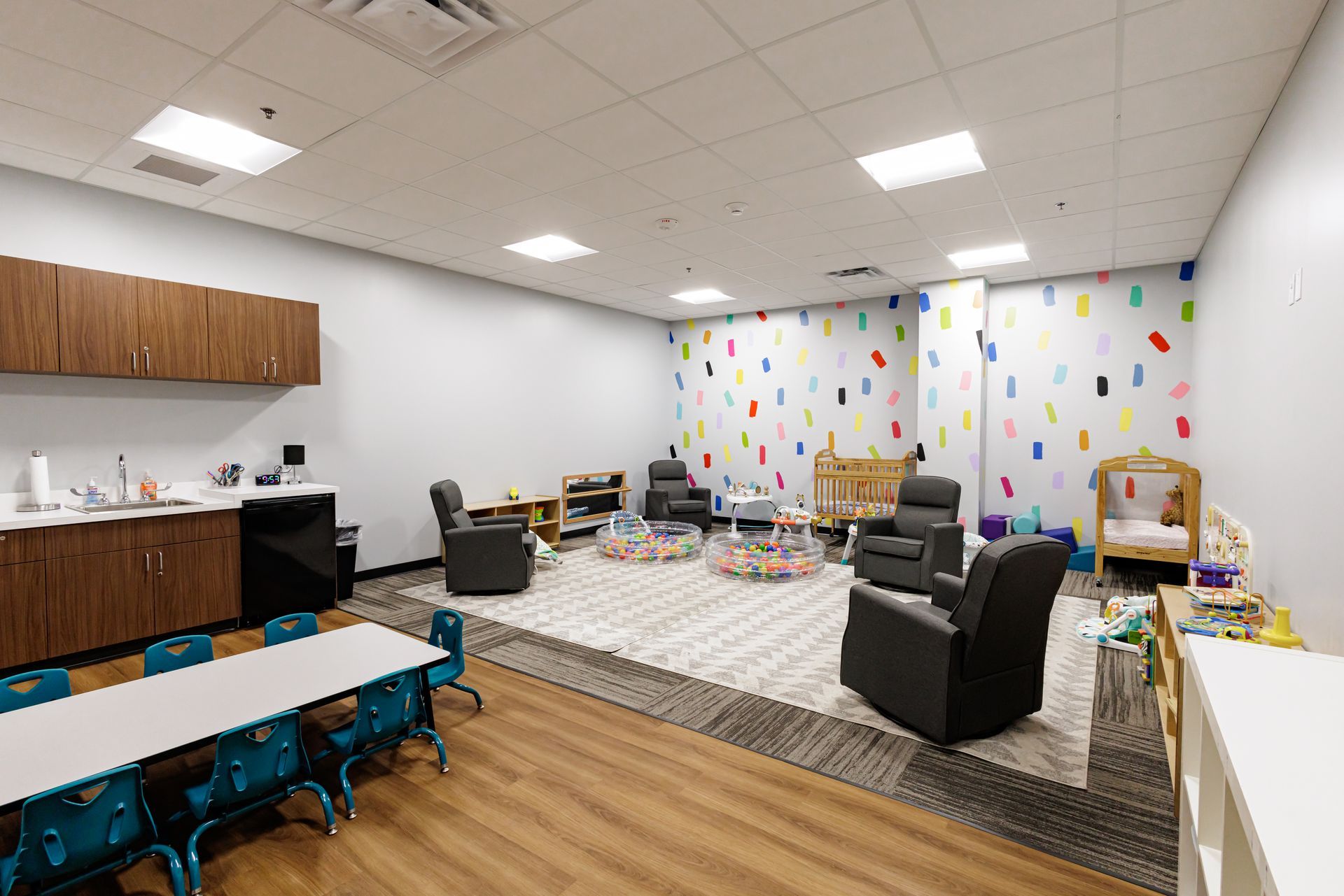 A nursery room with a sink area, small tables, chairs, reclining seats, play areas, and colorful wall decals.