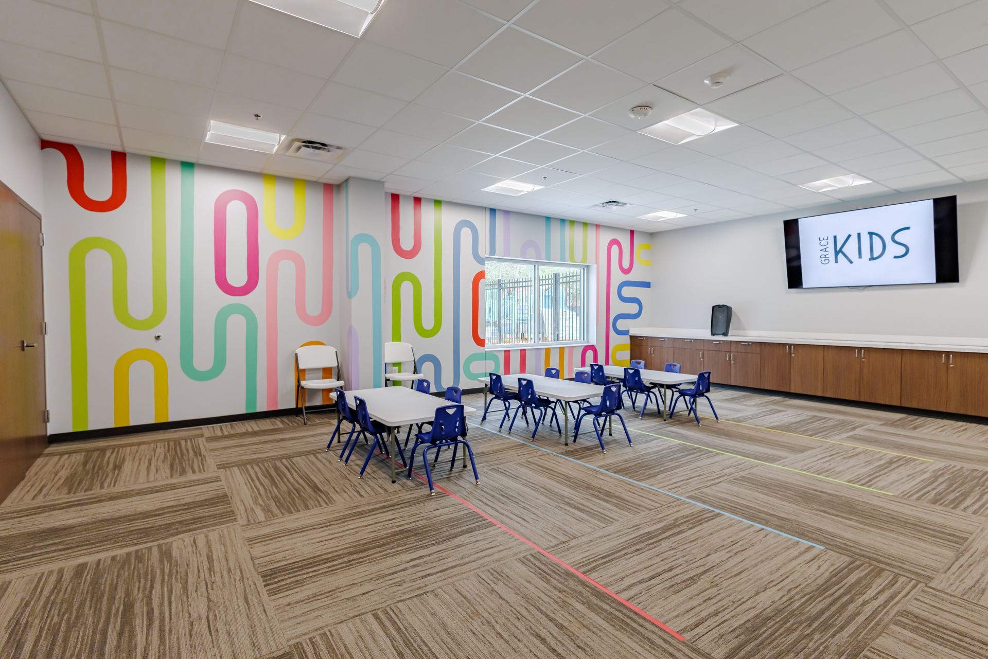 A bright, colorful kids' room with tables, small blue chairs, patterned carpet, and a wall-mounted TV displaying 