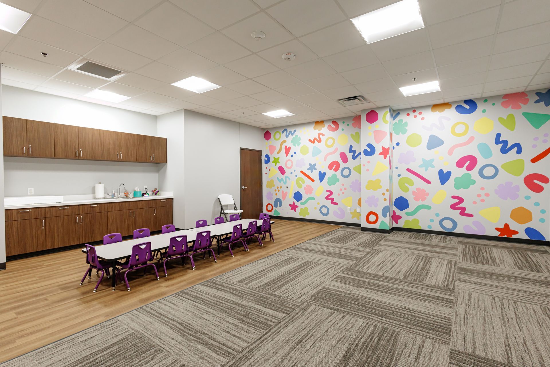 A colorful, patterned classroom with a long table, purple chairs, wooden cabinets, and gray patterned carpet.