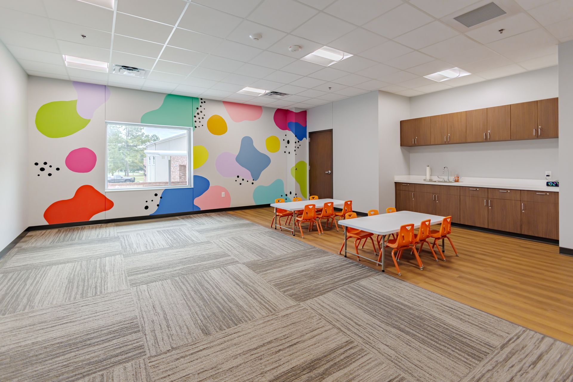 A bright, modern playroom with a colorful, abstract mural, tables with orange chairs, and wood cabinetry.