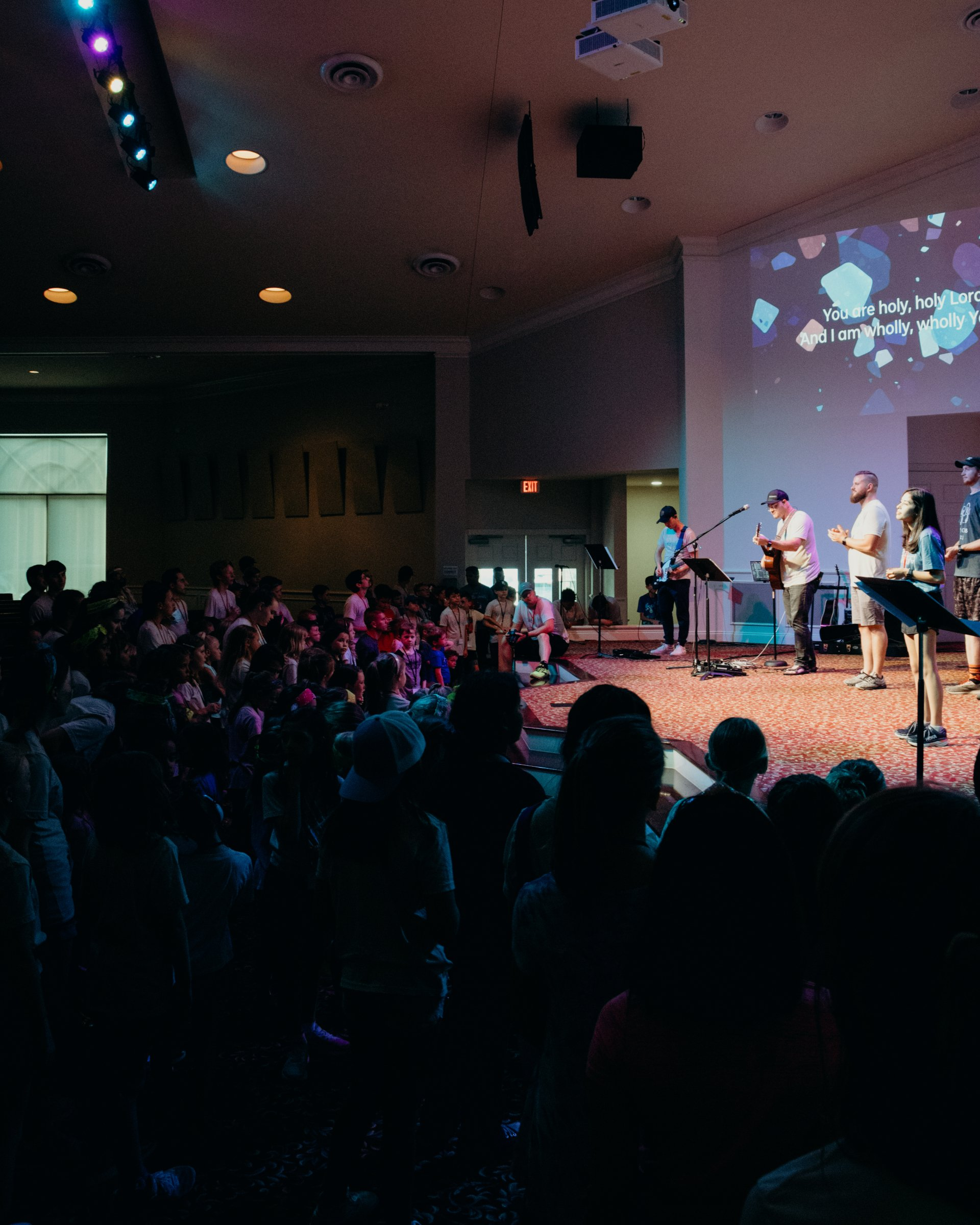 Worship band performing on stage in the GBC sanctuary, audience of children watching from below, projection on the wall.