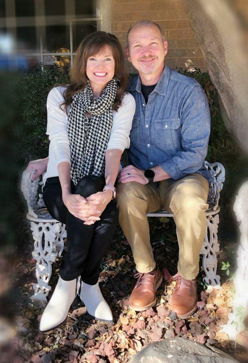 Couple sitting closely outdoors, smiling. Woman in scarf, white top, black pants. Man in denim shirt, tan pants.