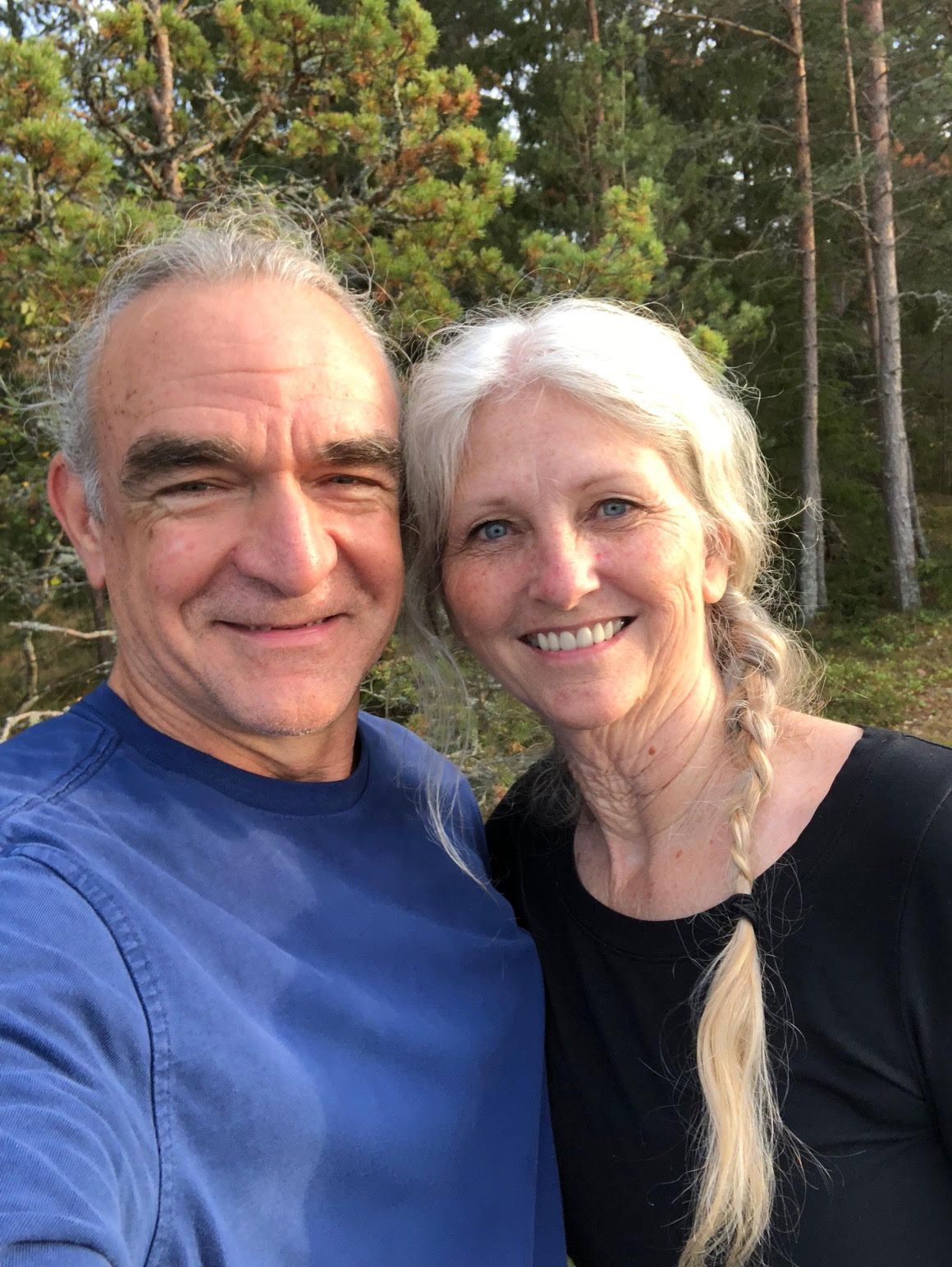 Couple smiling outdoors, man in blue shirt, woman with blonde braid. Green trees in background.