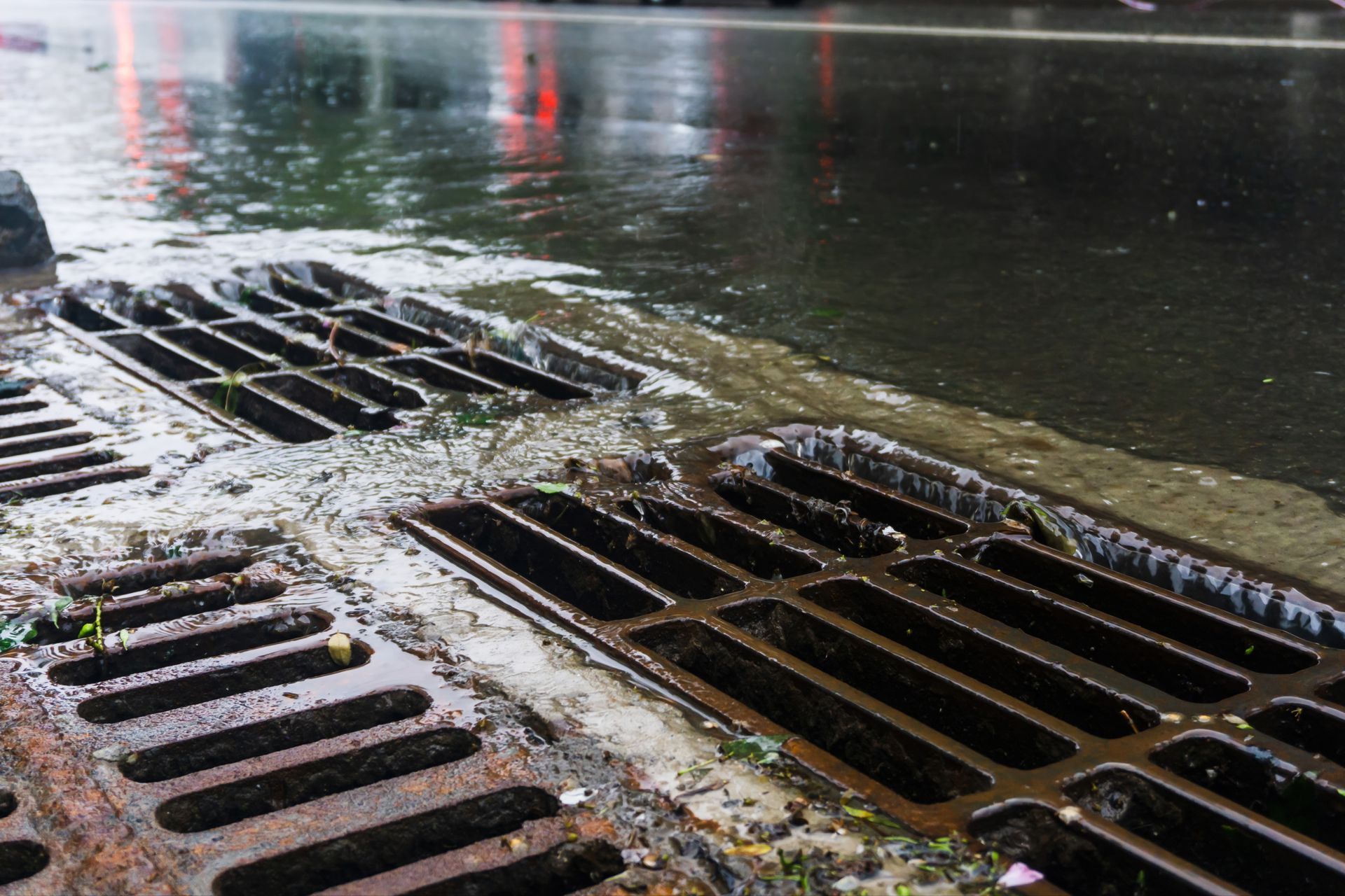 Photograph showing water flowing into rusty metal storm drains on a wet street surface. Photograph showing water flowing into rusty metal storm drains on a wet street surface.