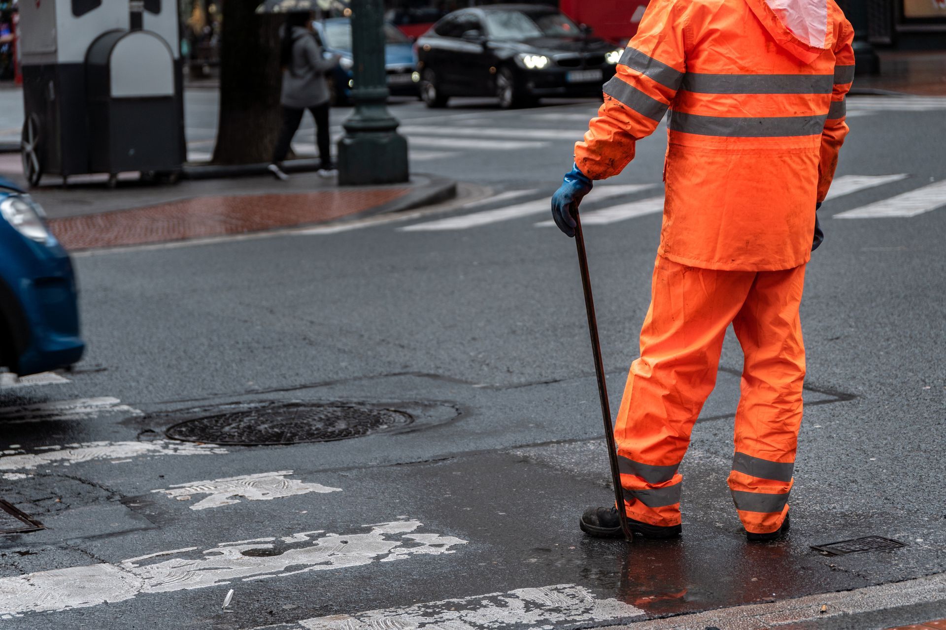 Worker in hi-vis gear performing storm drain maintenance on urban street in rainy conditions.