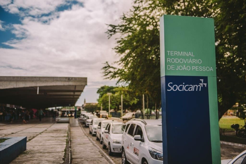 Placa do terminal rodoviário de João Pessoa, com uma fila de carros brancos, sob um céu nublado.