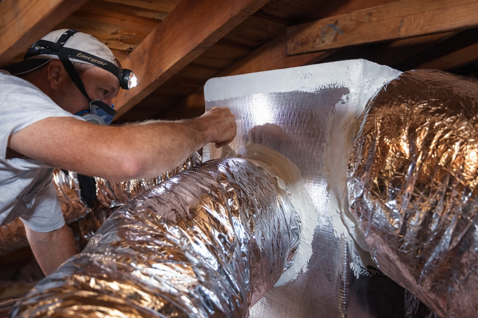 A worker wears a headlamp and respirator while sealing an HVAC duct connection with white mastic in an attic.