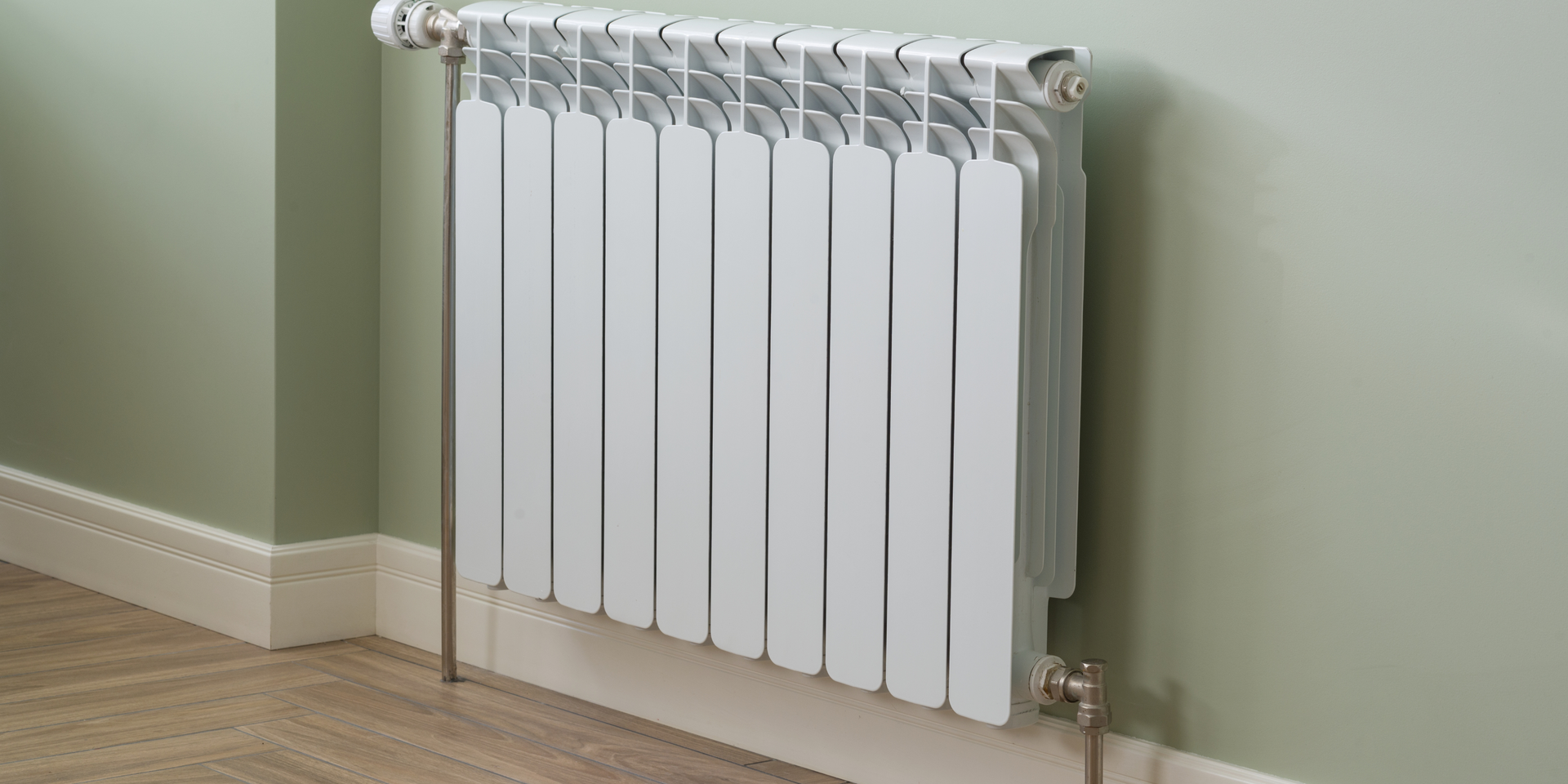 A white, multi-sectioned radiator mounted on a light green wall above a wooden floor.