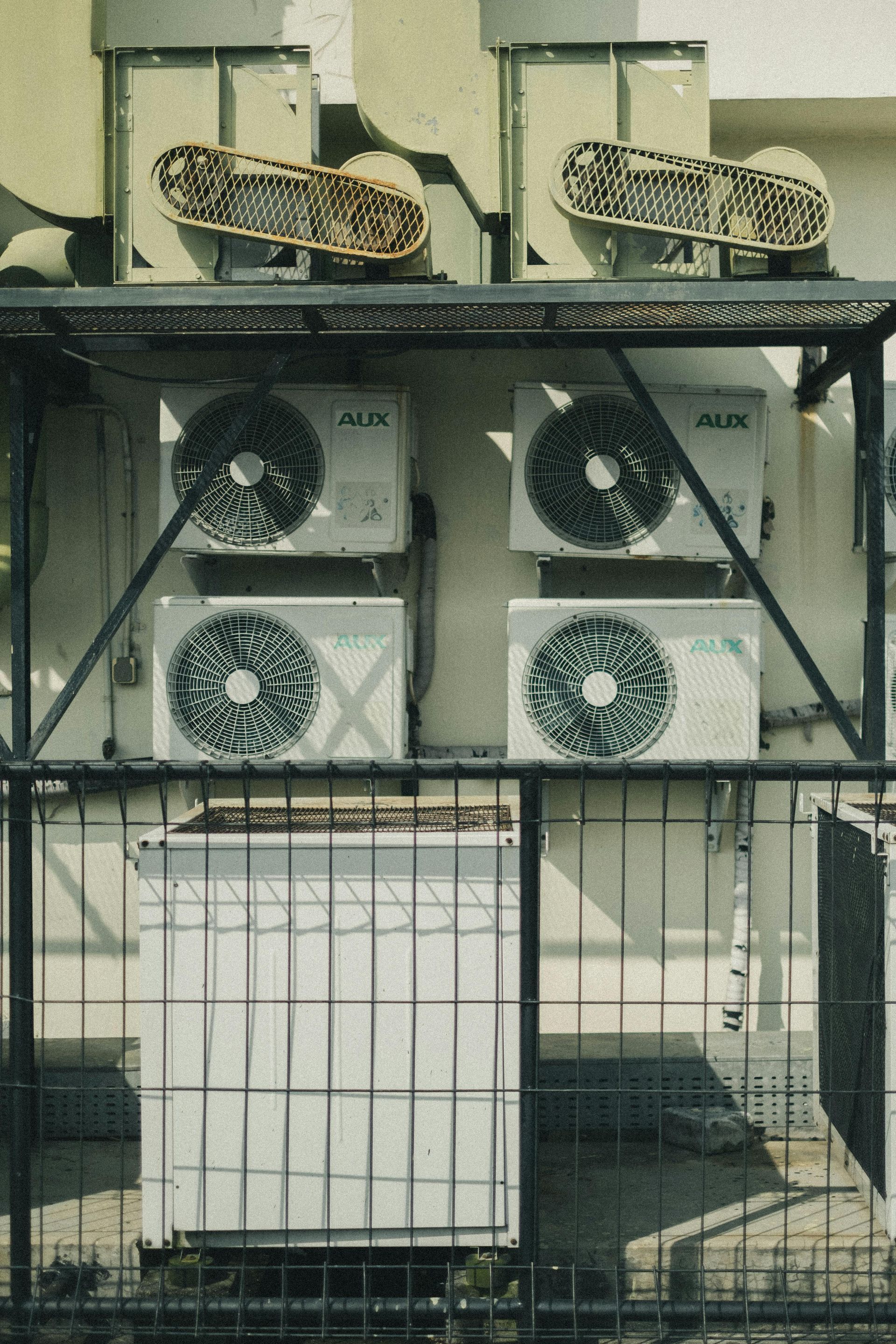 Stacked air conditioning units of various sizes and brands mounted on a wall behind a metal security fence.